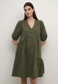 Olīvu zaļš, īsām piedurknēm, kleita ar V veida izgriezumu, savilkta krūšu daļa un izplešanās svārki. Izgatavota no viegla auduma ar maigu spīdumu.