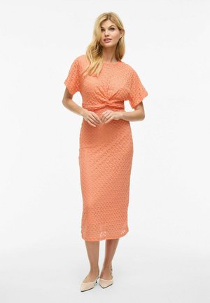 Peach textured midi dress with short sleeves, cinched waist, and a twisted front detail. Worn with light-colored heels.