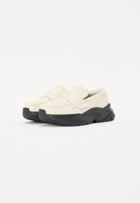 Des chaussures slip-on couleur crème avec un dessus texturé, dotées d'une semelle en caoutchouc noir épaisse et d'une large sangle sur le cou-de-pied pour le maintien.