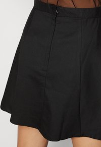 Falda negra con una tela suave, forma acampanada, pliegues frontales, detalle de cremallera oculta y un dobladillo que cae por encima de la rodilla.