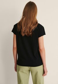 Černé tričko s krátkým rukávem, zaobleným lemem a volným střihem, v kombinaci se světle zelenými kalhotami. Hladká textilie, žádné viditelné vzory.