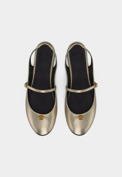 Χρυσά μεταλλικά φλατ παπούτσια με λουριά στον αστράγαλο και διακοσμήσεις κεφαλής Μέδουσας στην μύτη, σε συνδυασμό με μαύρα πέλματα.
