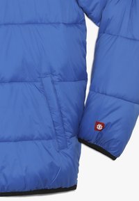 Veste matelassée bleue avec une texture lisse et brillante. Comprend une poche latérale et un écusson logo rouge sur la manche. Poignets élastiques pour un ajustement parfait.