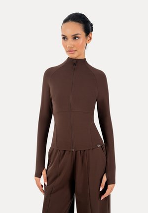 Mujer con una camiseta marrón ajustada de manga larga con cremallera y agujeros para el pulgar, y pantalones marrones sueltos a juego sobre un fondo liso.