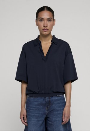 Frau mit zurückgebundenem dunklen Haar, die ein lockeres marineblaues Kurzarmshirt mit Kragen und blaue Jeans trägt, steht vor einem einfarbigen Hintergrund.