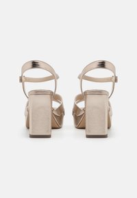 Sandalias de tacón alto beige con un acabado metálico, tacones de bloque, correas delgadas en los tobillos y una superficie texturizada, con un diseño entrecruzado.