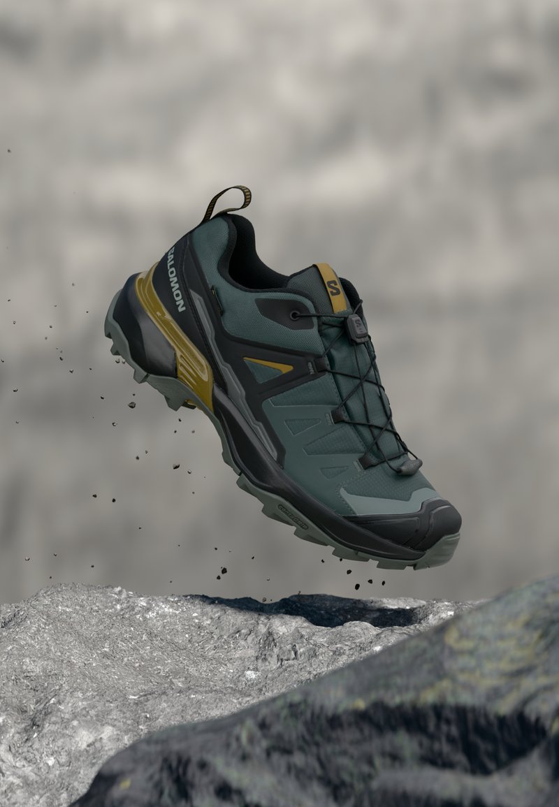 Trailrunningschoen in groen en zwart met gele accenten, zwevend boven rotsachtig terrein met kleine vuildeeltjes die vallen.