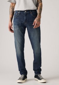 Dark blue denim jeans with light fading. Straight fit, five-pocket design, visible stitching, and metal button closure. Worn with black canvas sneakers.