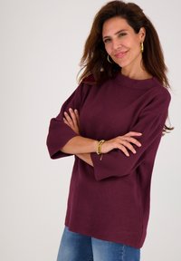 Vrouw met lang bruin haar, gekleed in een bordeauxrode trui, blauwe jeans, gouden oorringen en armbanden, die met gekruiste armen tegen een witte achtergrond staat.