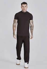 Dunkelbraunes, tailliertes T-Shirt mit kurzen Ärmeln, kombiniert mit schwarzen, gerippten Hosen. Das Outfit wird durch weiße, lässige Turnschuhe abgerundet.