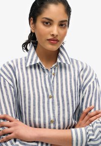 Camisa de botones a rayas en azul y blanco, hecha de una tela ligera. Presenta un cuello clásico y botones visibles en el frente.