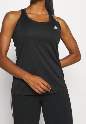Femme portant un haut de sport noir sans manches Adidas et des leggings noirs avec des bandes blanches, debout avec une main sur l'abdomen.