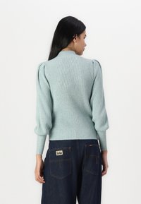 Light blue ribbed sweater with puffed sleeves, high collar, and fitted waist, styled with dark denim pants featuring a logo patch.