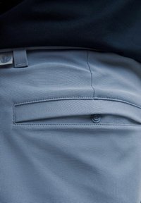 Heledad sinised püksid, millel on sile tekstuur, tagaküljel nupuga tasku ja õmmeldud detailid.