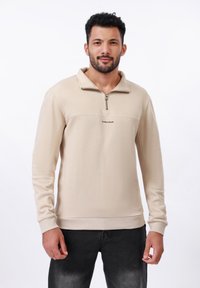 Sweatshirt zippé beige en tissu doux, avec un panneau horizontal contrastant, des poignets côtelés et un logo au centre.