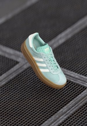 Jasnoniebieski sneaker Adidas Gazelle z białymi paskami i gumową podeszwą, unoszący się nad szarym metalowym rusztem w tle.
