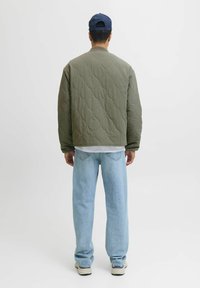 Gequilted olijfgroene jas met een ronde hals, gedragen over een wit shirt, gecombineerd met lichtblauwe jeans en beige sneakers.