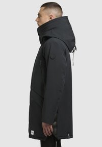 Jachetă parka neagră cu guler înalt, glugă texturată, buzunar cu fermoar și fante laterale, având un patch cu logo pe mânecă.