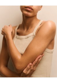 Débardeur beige avec un décolleté carré, doté de fines bretelles. Un tissu lisse recouvre la peau exposée sur les bras, mettant en valeur la texture naturelle de la peau.