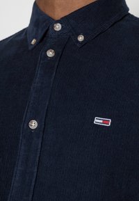 Námořnická manšestrová košile s límečkem na knoflíky, vybavená průhlednými knoflíky a malým vyšitým logem s červenými, bílými a modrými akcenty.