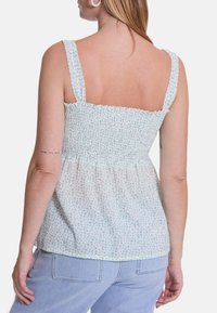 Helles grünes Oberteil mit floralem Druck, gesmoktem Rücken und breiten Trägern. Weicher, leichter Stoff und entspannter Schnitt. Kombiniert mit hellblauen Jeans.