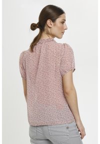 Donna con i capelli castani raccolti in un bun, che indossa una blusa floreale rosa a maniche corte e pantaloni grigio chiaro, in posizione laterale su uno sfondo bianco.