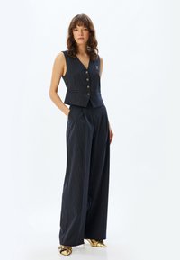 Gilet rayé bleu marine avec boutons dorés et coupe ajustée, accompagné de pantalons larges assortis. La texture semble lisse et structurée.