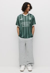 Grøn stribet fodboldtrøje med hvid krave og detaljer, der har teksten "ASSOCIATION D'ÉQUIPE", parret med grå sweatpants og hvide sneakers.