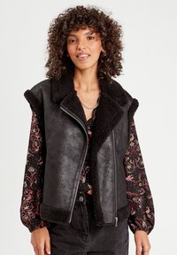 Gilet in finta pelliccia nera con alto collo e chiusura a zip. Sembra morbido con una finitura testurizzata. Indossato sopra una blusa a maniche lunghe con motivo.