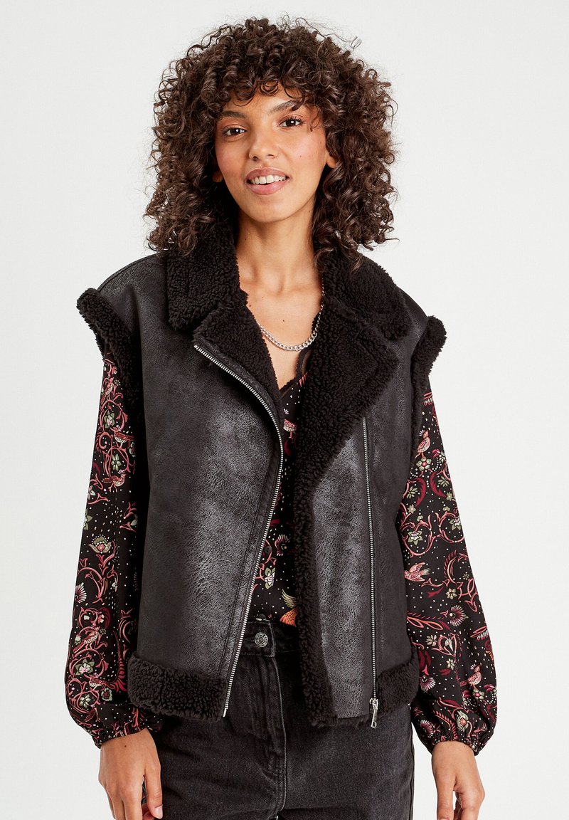 Gilet in finta pelliccia nera con alto collo e chiusura a zip. Sembra morbido con una finitura testurizzata. Indossato sopra una blusa a maniche lunghe con motivo.