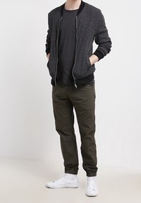 Pánská bomber bunda v tmavě šedé texturované tkanině s černými detaily, kombinovaná s tmavým tričkem a olivově zelenými zúženými kalhotami. Bílá sneakers dotváří celý vzhled.