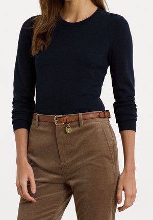 Mujer con una camiseta negra ajustada de manga larga metida dentro de unos pantalones de pana marrón de cintura alta, con un cinturón de cuero marrón y una hebilla dorada decorativa.