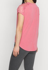 T-shirt rosa a maniche corte con dettagli in pizzo sulle spalle, materiale morbido, orlo arrotondato e vestibilità rilassata, abbinato a jeans grigi.