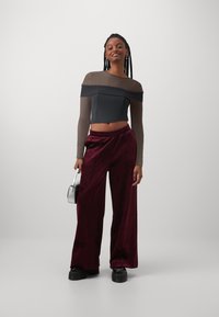 Schwarzes und braunes schulterfreies Crop-Top mit langen durchsichtigen Ärmeln, kombiniert mit weiten burgunderfarbenen Samt-Hosen. Enthält eine silberne Handtasche.