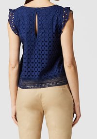 Haut bleu marine avec détails en dentelle, manches courtes à volants et dos en forme de trou de serrure. L'ourlet présente un motif décoratif en dentelle. Associé à un pantalon beige.