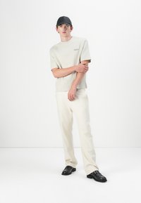 Beige T-Shirt mit kurzen Ärmeln und dezentem Logo, kombiniert mit cremefarbenen Hosen. Eine schwarze Mütze und Schuhe runden den Look ab. Glatte Stofftextur.