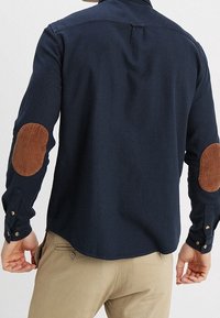 Marinblå långärmad skjorta med bruna manchesterelbow-patchar, knappmanchetter och en något rundad nederkant. Slät tygtextur.