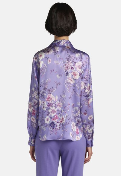 Woman with short dark hair wearing a long-sleeve purple floral blouse and solid purple pants, standing with back facing the viewer.