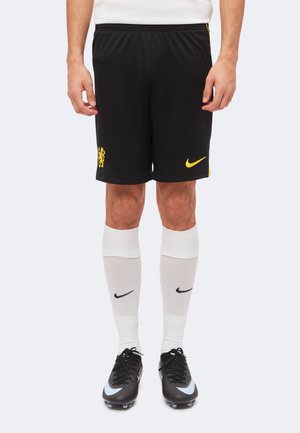 Hombre con pantalones cortos deportivos negros con logo de Nike amarillo, calcetas blancas hasta la rodilla con el símbolo negro de Nike y zapatos de fútbol negros.