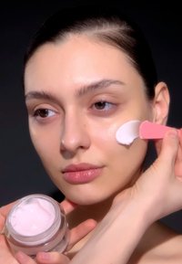 A glass jar with pink cream and silver lid, with a hand applying the cream to a face using a pink spatula; skin is smooth and glowing.
