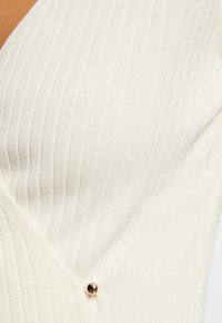 Gros plan sur un tissu côtelé crème en tricot avec un petit bouton rond doré sur un vêtement ajusté.