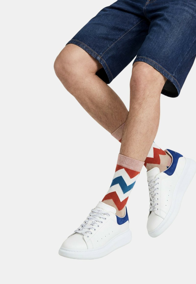 Beine übereinandergeschlagen, wearing Jeansshorts, beige-rot-blaue gesprenkelte Socken und weiße Sneakers mit blauen Akzenten.