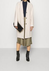 Ljusbeige oversized kappa med knappstängning, marinblå topp, olivgrön silkjurk och svarta ankelstövlar. Väska med kedjedetalj.