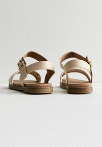 Sandale beige avec deux sangles réglables, semelle plate et détails de couture minimalistes. Fabriquée en matériau synthétique avec une texture lisse.
