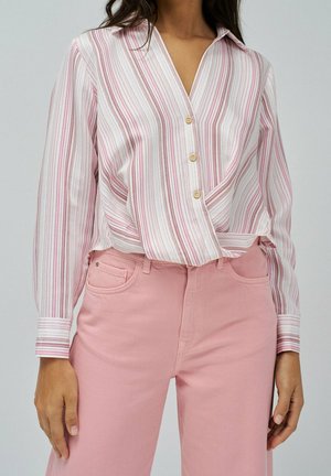 Femme portant un chemisier à boutons rayé verticalement blanc et rose, rentré dans un pantalon taille haute rose pastel, debout les bras détendus.