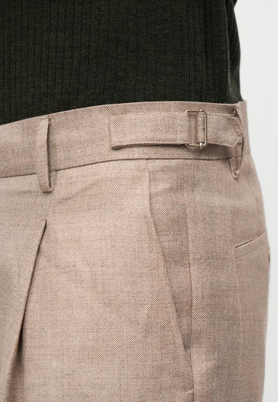 Κοντινό πλάνο μπεζ λινών παντελονιών με ρυθμιζόμενο αγκράφα στο πλάι και θηλιά για ζώνη, φορεμένα πάνω από σκούρο πράσινο ανάγλυφο τοπ.