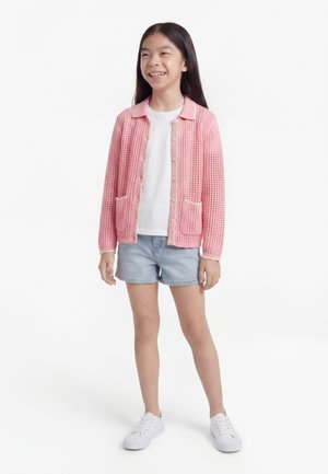 Chica de pie y sonriendo, lleva un cárdigan de punto rosa, camiseta blanca, pantalones cortos de mezclilla y zapatillas blancas sobre un fondo blanco.