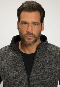 Chaqueta de cremallera de color gris oscuro con un patrón texturizado y un cuello alto, superpuesta a una camiseta negra. La tela parece suave y duradera.
