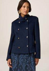 Marineblaue doppelt-knopfte Jacke aus strukturiertem Stoff, mit goldenen Knöpfen, hohem Kragen und tailliertem Schnitt.