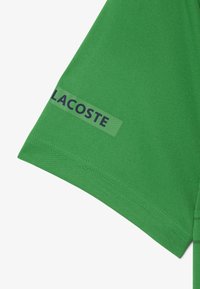 Manica in tessuto verde con orlo cucito e un'etichetta trasparente che mostra il nome del marchio "LACOSTE" in lettere grasse.